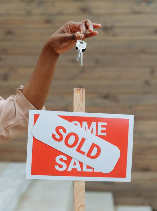 Hand holding house keys over a 'sold' sign representing a real estate transaction completed by an investor