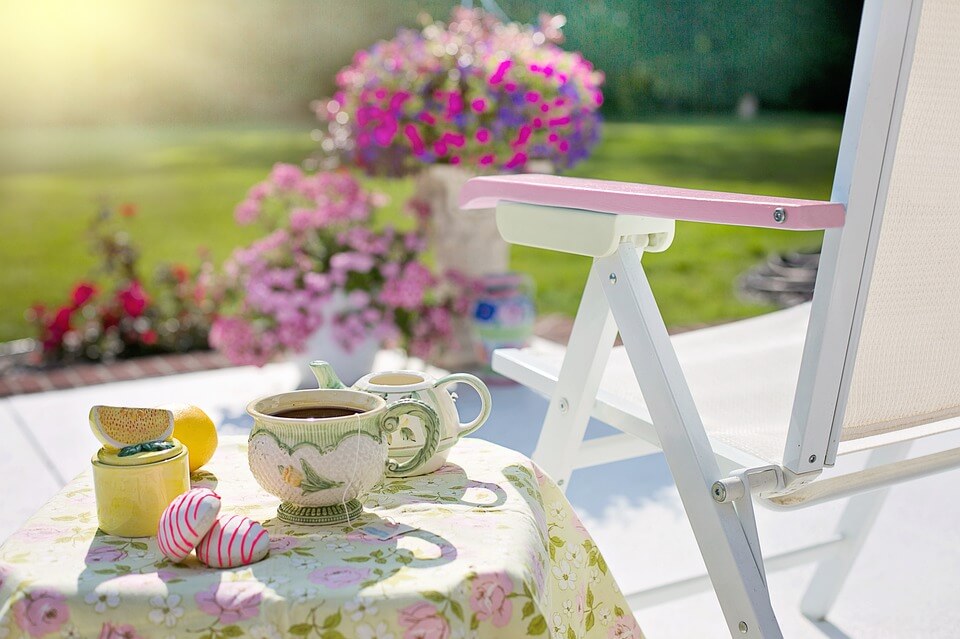 Porch table set with tea cups and cookies alongside a chair surrounded by abundant fresh flowers creating an inviting al fresco dining setup