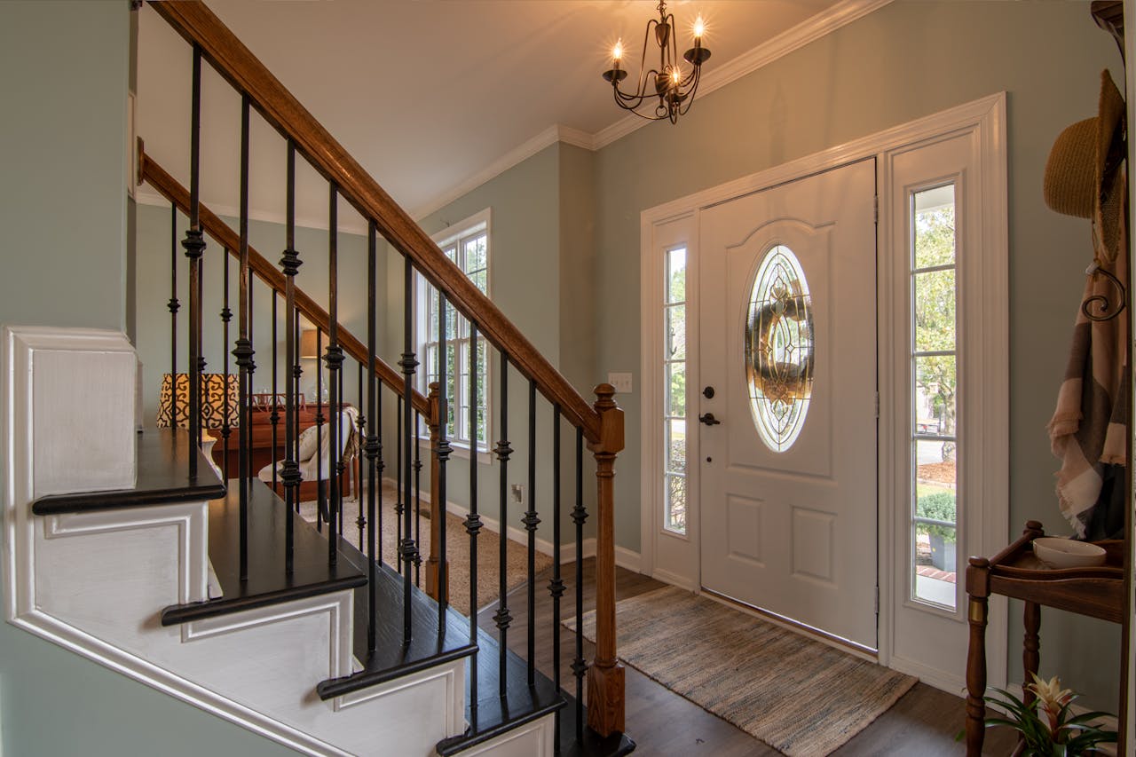 Wooden staricase, brass chandelier, large white front door. Image by Pexels